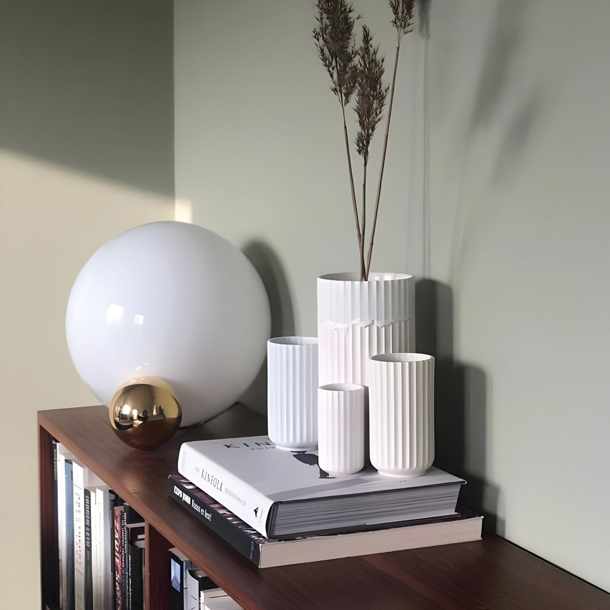 A modern shelf with several books, white ribbed vases with dry stems, and a Soul Table Lamp - Copper, all set against a light green wall for a sleek modern minimalist look.