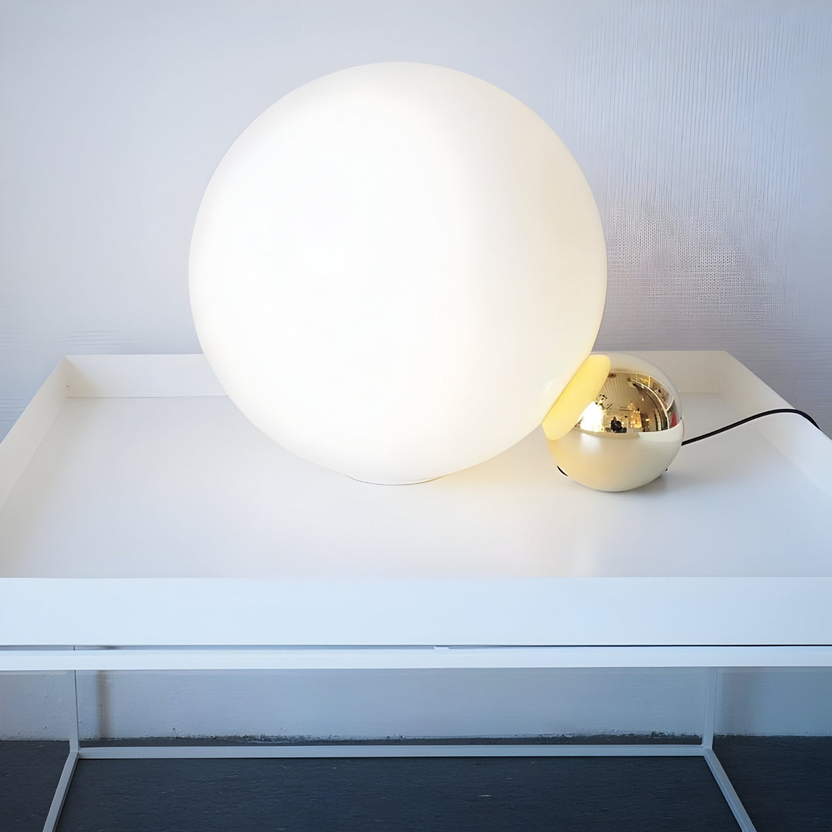 The Soul Table Lamp - Gold features a glowing white glass shade paired with a smaller reflective gold sphere, displayed on a minimalist white glass-top table. The lamp’s cord is visible on the right.