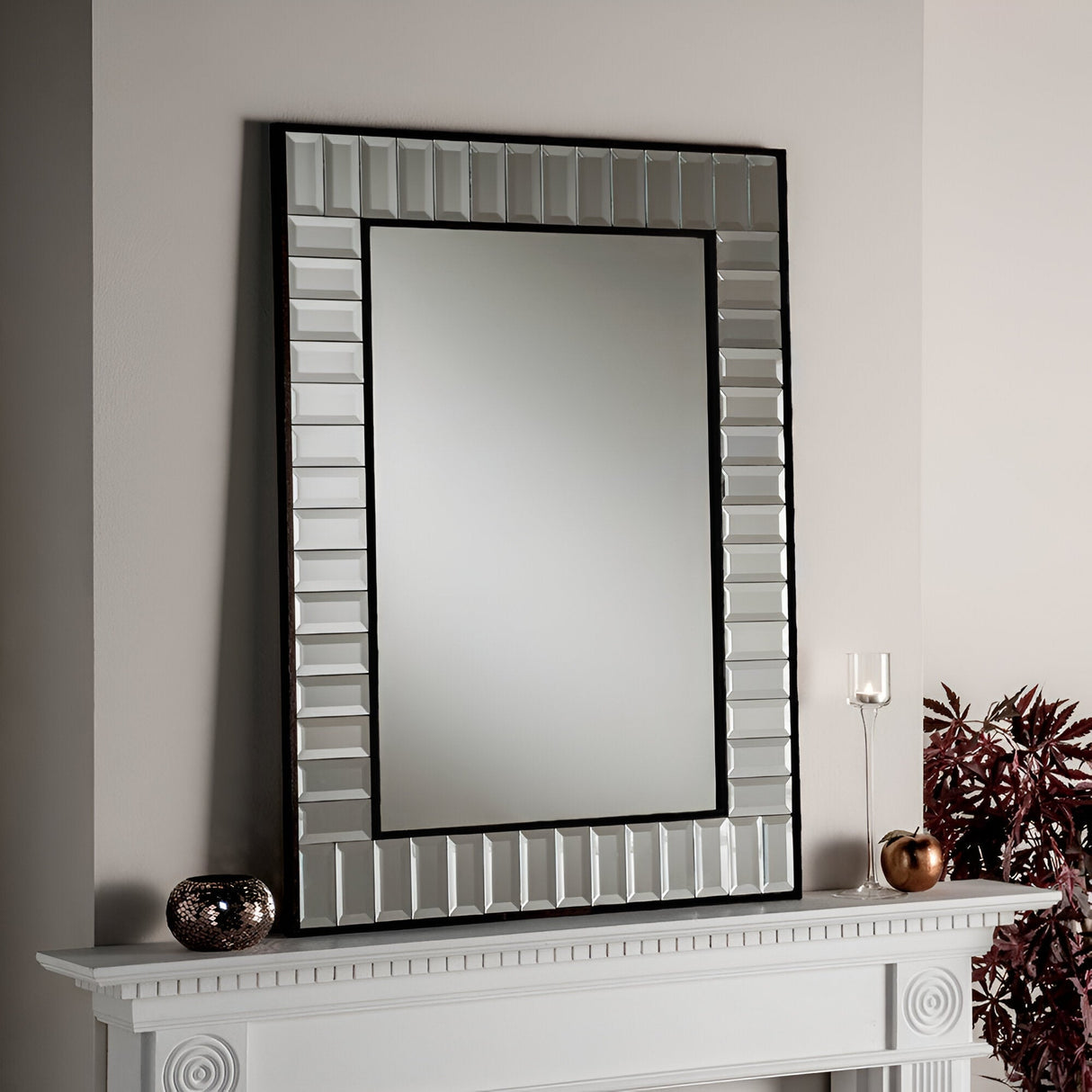 The Brillix Rectangle Wall Mirror in black, featuring a textured geometric border, hangs above a white fireplace mantel. On the mantel are decorative items, including a copper-colored spherical object and a transparent candle holder. A plant with dark leaves on the right enhances the modern design.
