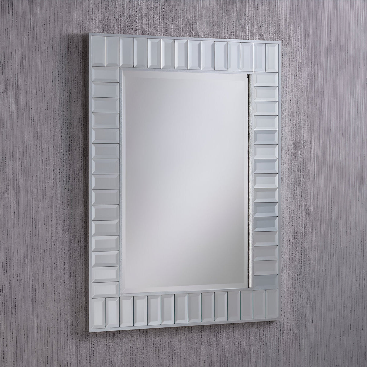 The Brillix Rectangle Wall Mirror - Silver, ideal for those passionate about interior design, features a silver frame adorned with a pattern of raised, alternating rectangles. When mounted on a textured grey wall, it beautifully reflects soft, neutral light, enhancing the ambiance of the space.