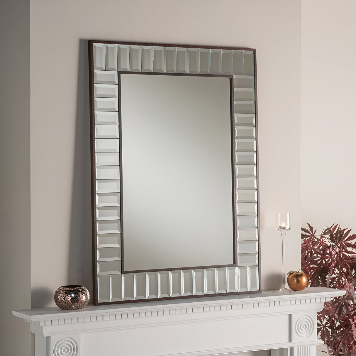 The Brillix Rectangle Wall Mirror, featuring a silver and wood-textured frame, hangs prominently above a white fireplace mantel. Adorning the mantel are a small copper vase, a glass candle holder, and a potted plant with red leaves, all of which enhance the charm of any living space.