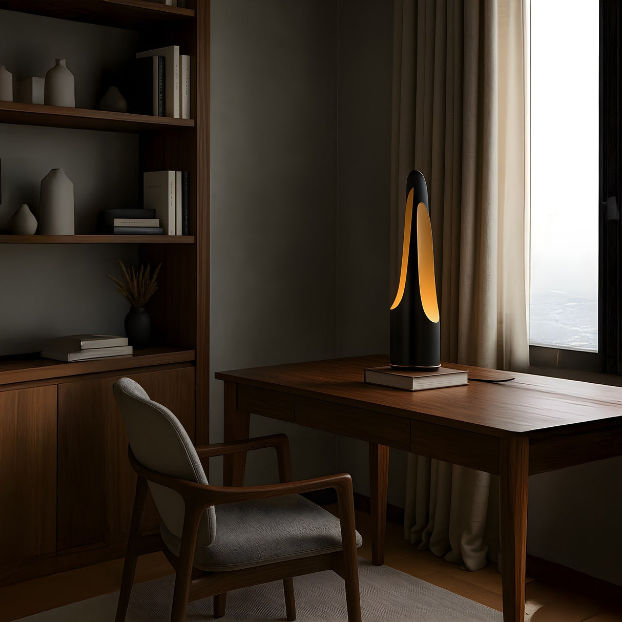 A modern home office with a wooden desk, cushioned chair, built-in shelves displaying vases, and the Stealth Table Lamp in Brass & Black glowing warmly beside a window with beige curtains.