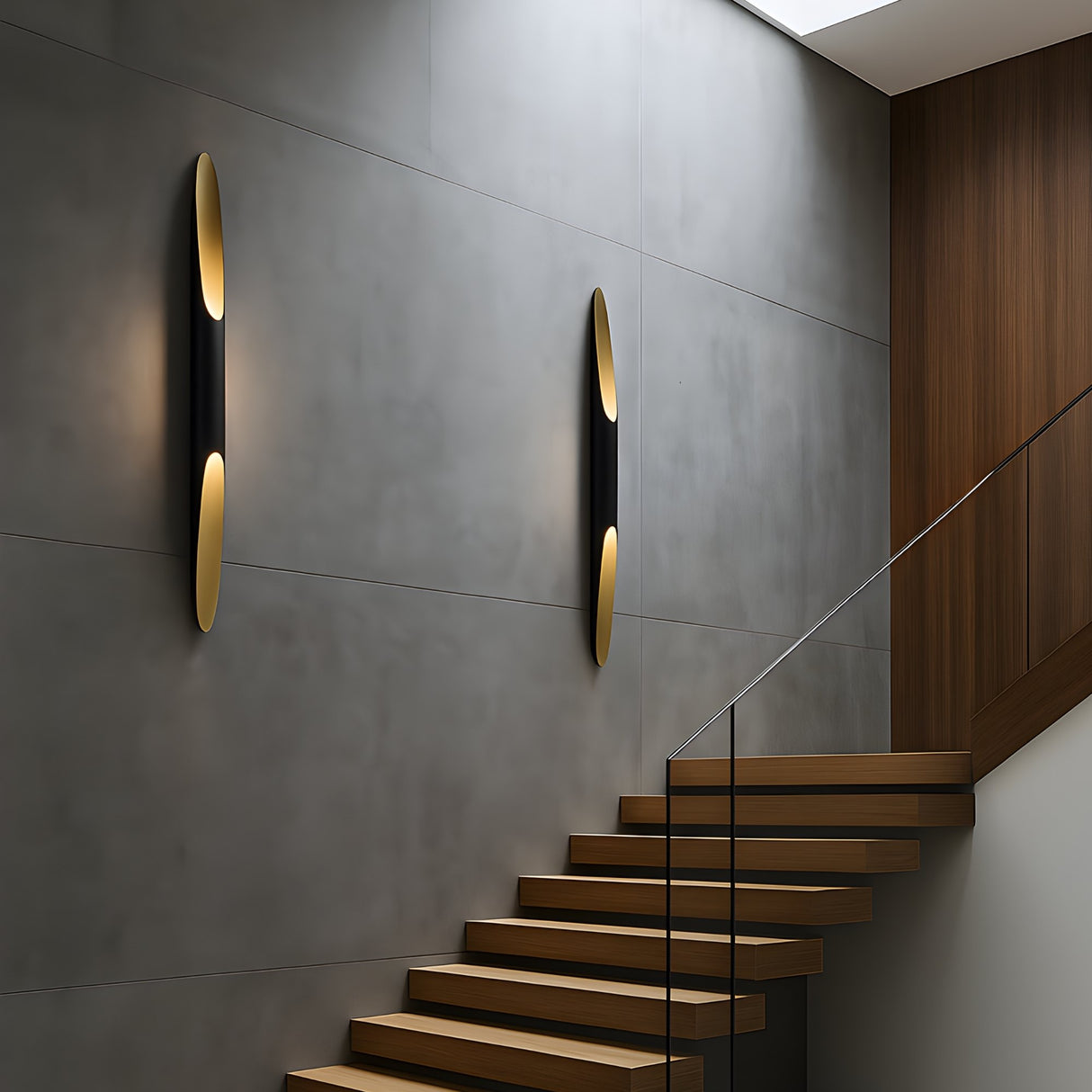 A modern staircase with wooden steps and glass railing rises beside a gray concrete wall, which is accented by two Stealth 2 Light Wall Lights in brass and black emitting a soft glow.