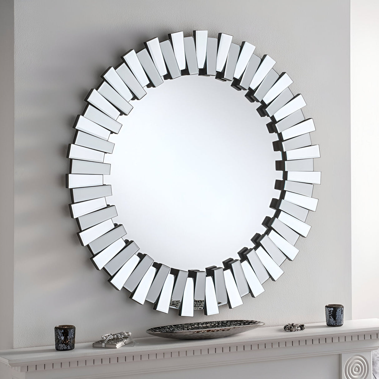 A large Stella Circle Wall Mirror with a round sunburst design is mounted on the white wall above a mantel. The frame showcases intricate detailing with angular mirrored panels. Unique decorative elements adorn the mantel, including small vases and accents.