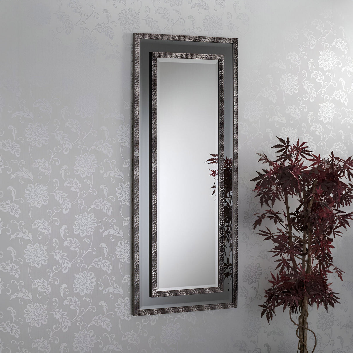 The Speciax Rectangle Wall Mirror - Grey adorns the patterned wall with elegance. To its right, a plant with burgundy leaves introduces a natural element that complements the modern decor and enhances the minimalist atmosphere.