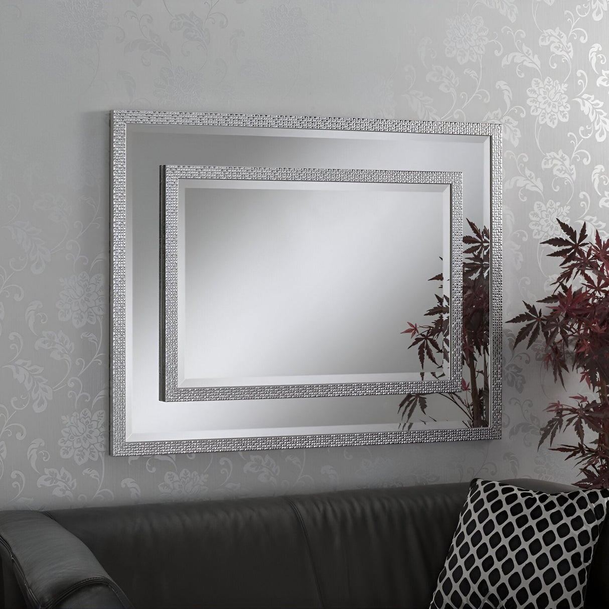 A Speciax Rectangle Wall Mirror - Silver with an intricate border graces the wallpapered wall, reflecting the room's chic vibe. The dark couch beneath it boasts a decorative pillow with a geometric pattern, while a nearby plant adds an organic touch to this stunning home decor scene.