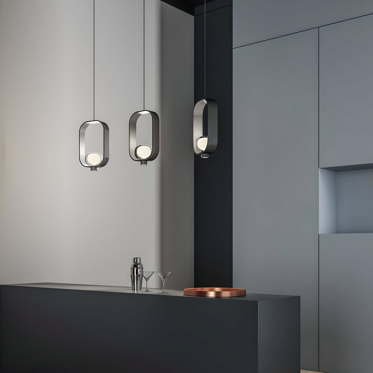 A modern kitchen with matte gray cabinets and a dark island features three Strappa Suspension Lights in gold & black. On the island are a cocktail shaker, two martini glasses, and a round copper tray.