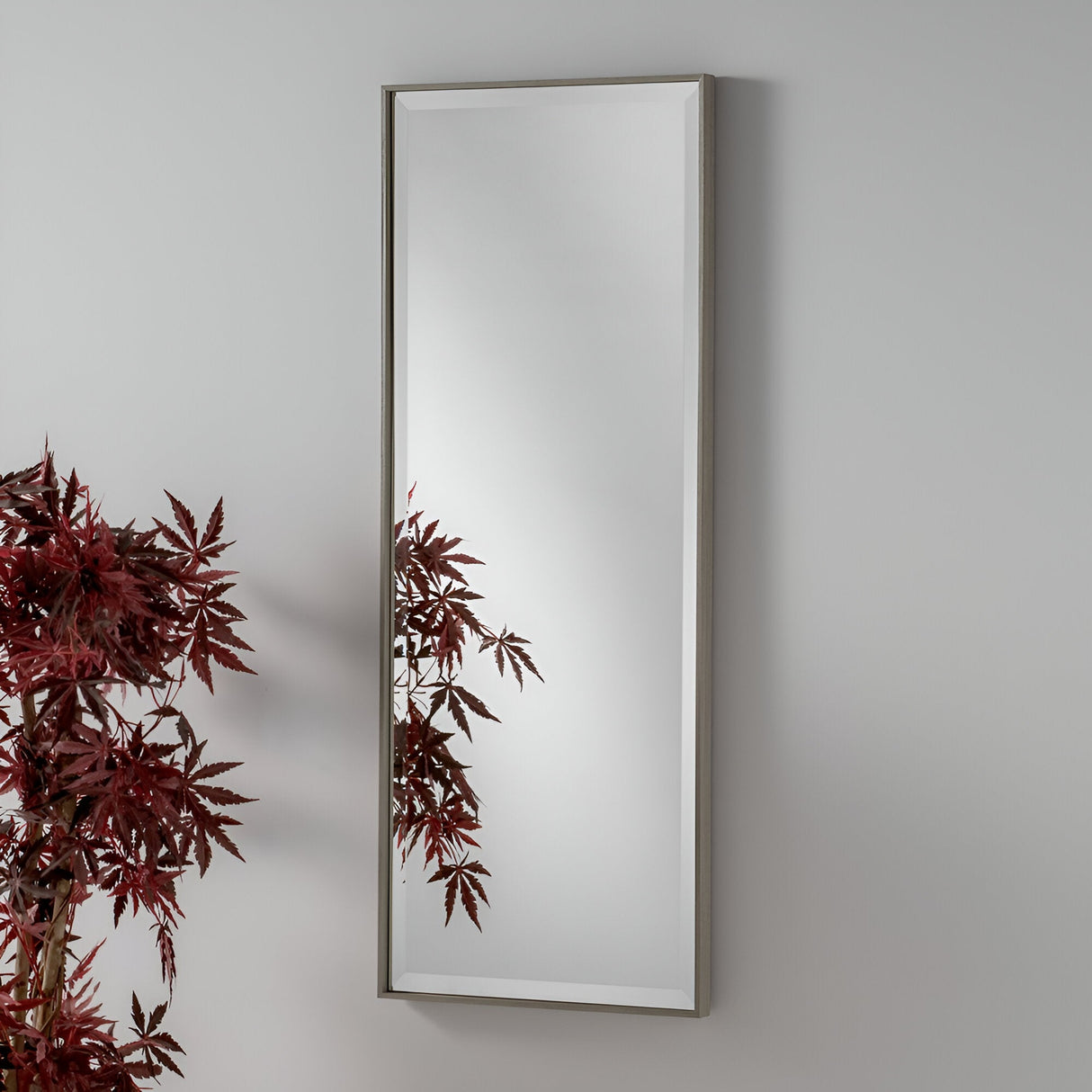 An Aetheris Rectangle Wall Mirror - Grey with a thin frame is mounted on a light gray wall. In the foreground, a plant with burgundy leaves is visible, partially reflected in this minimalist design element of home decor.