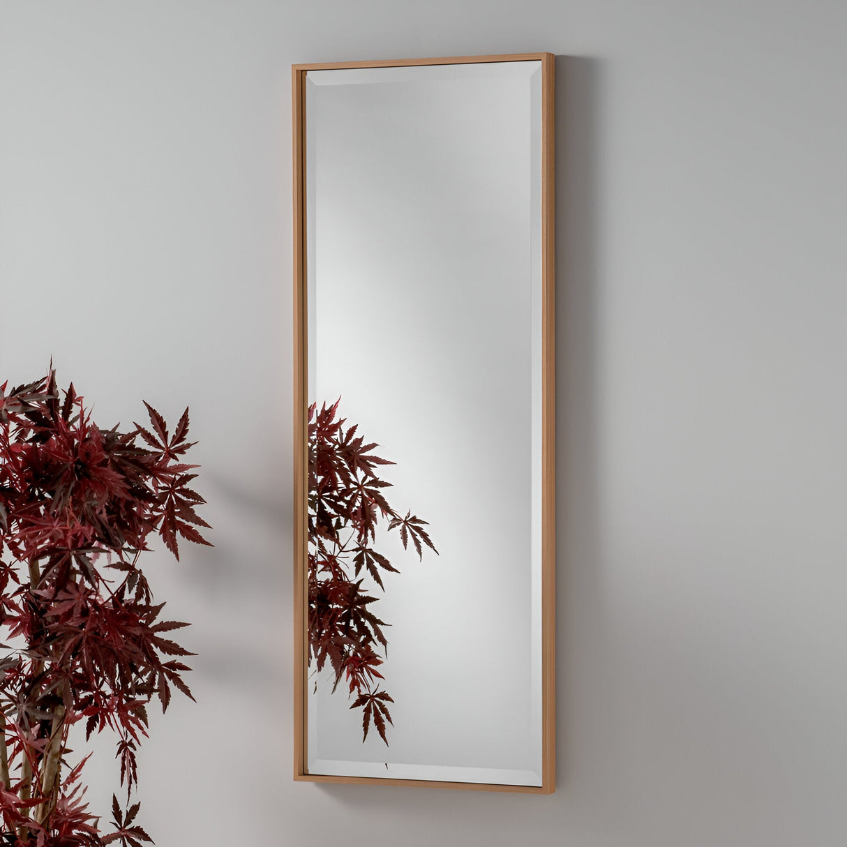 The Aetheris Rectangle Wall Mirror - Oak, with its tall, rectangular design and wooden frame, is mounted on a light gray wall. It elegantly reflects the leaves of a nearby red maple plant on the left side, seamlessly blending rustic charm with a modern aesthetic.