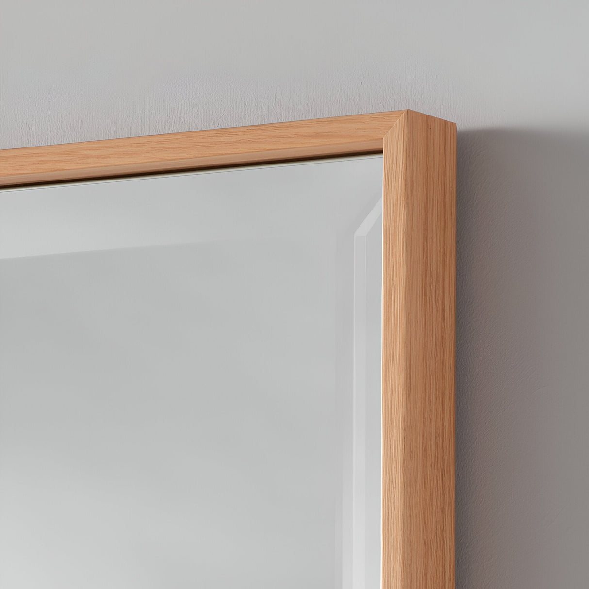 The Aetheris Rectangle Wall Mirror - Oak features a close-up view of its wooden frame, exuding rustic charm. The frame's smooth, natural finish beautifully complements the reflective surface, capturing the subtle gray wall and ambient light to create a minimalist yet modern aesthetic.