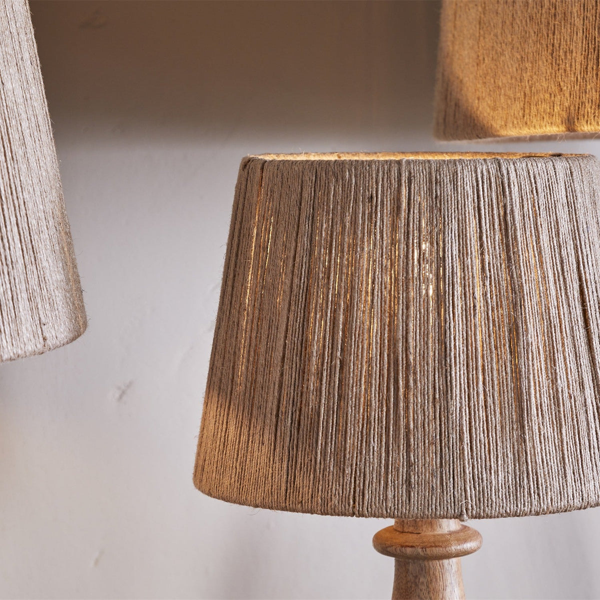 Tiwa Extra Large Woven Jute Lampshade in natural, a tapered drum of closely wound jute cord on a wooden lamp base, shown beside similar shades in a softly lit room.