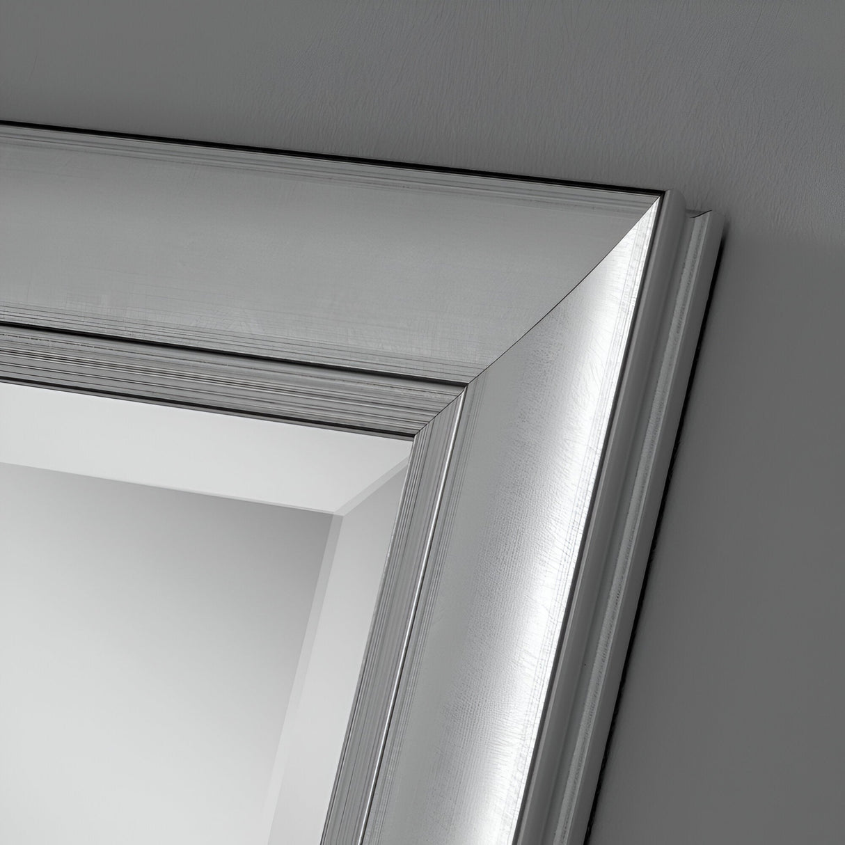 A close-up view of a corner of the Tivoli Rectangle Wall Mirror - Silver, showcasing its silver frame with a brushed aluminum finish set against a gray wall. The frame exhibits clean lines, bringing a modern touch to any space.
