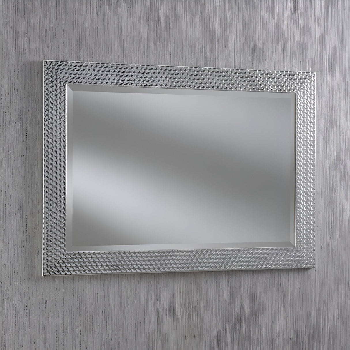 The Glimira Rectangle Wall Mirror - Silver features a textured, geometric-patterned frame and is mounted on a light gray wall, enhancing natural light while showcasing its sleek, modern design.