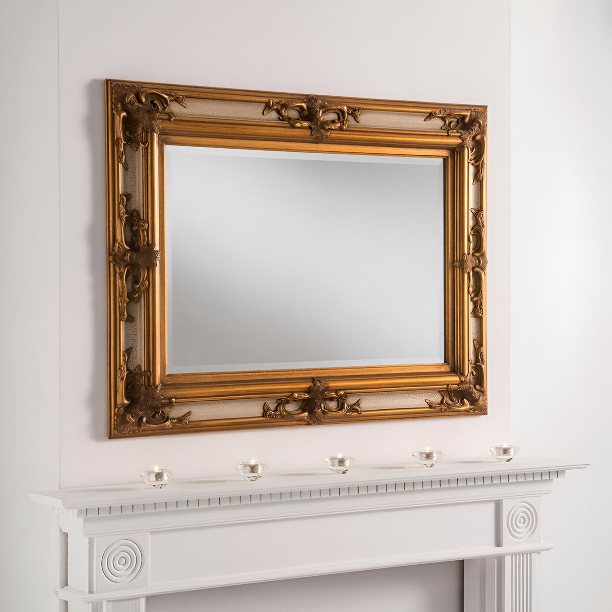 The Lucet Rectangle Wall Mirror in Gold/Ivory radiates timeless luxury as it hangs above the white mantelpiece. Five small glass tealight holders are evenly distributed across the mantel, which boasts decorative molding, further accentuating the mirror's elegant presence.