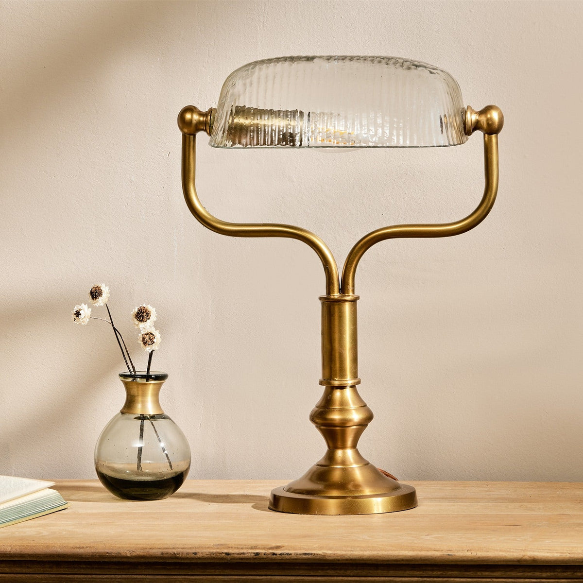 Ulani Vintage Desk Lamp in antique brass with a ribbed clear glass dome shade, twin-stem yoke and stepped round base, shown on a wooden surface beside a small glass vase.