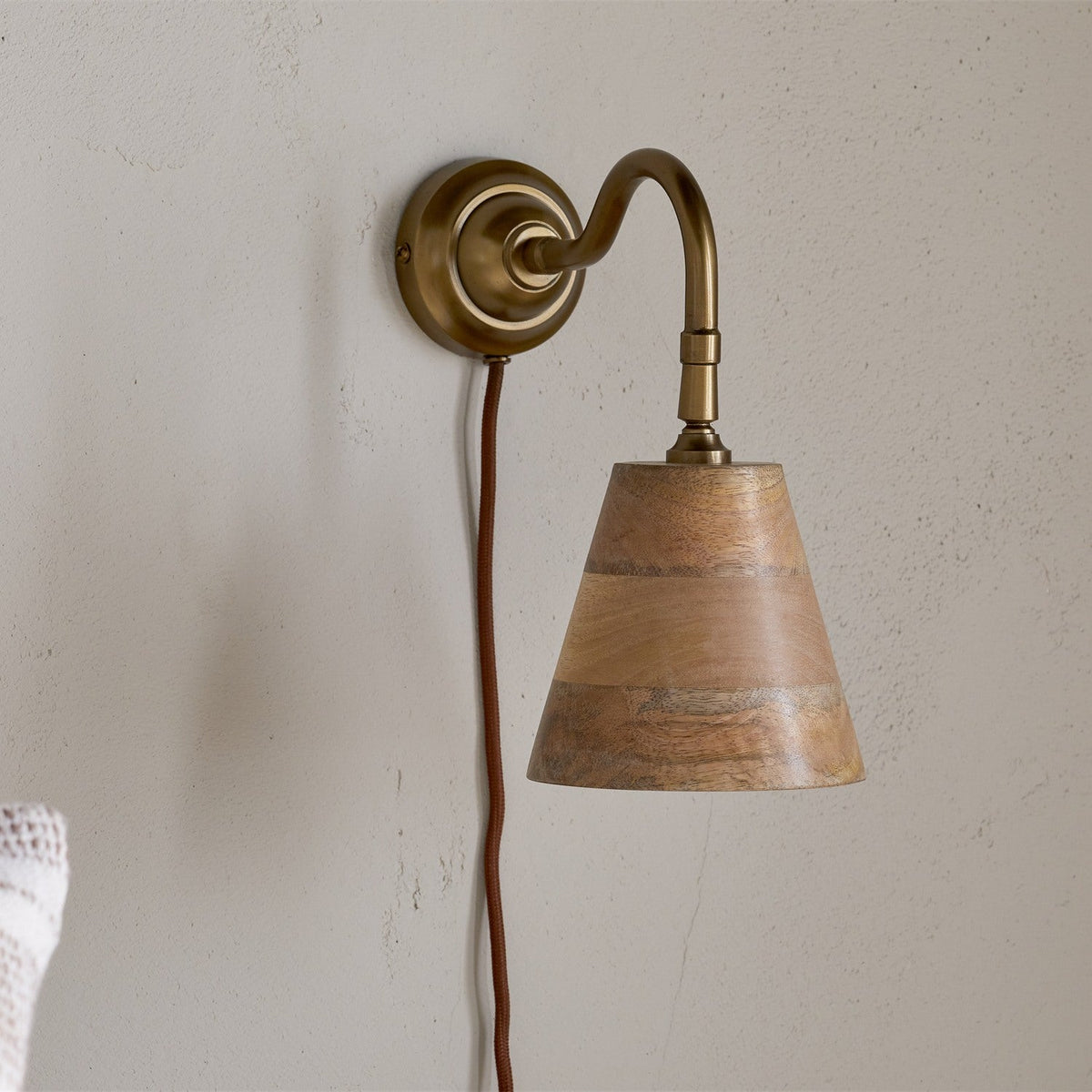 Vattu Mango Wood Plug In Wall Light in Natural, featuring an antique brass wall plate and swan neck arm with a mango wood cone shade and rust-coloured cord, shown against a textured light wall.