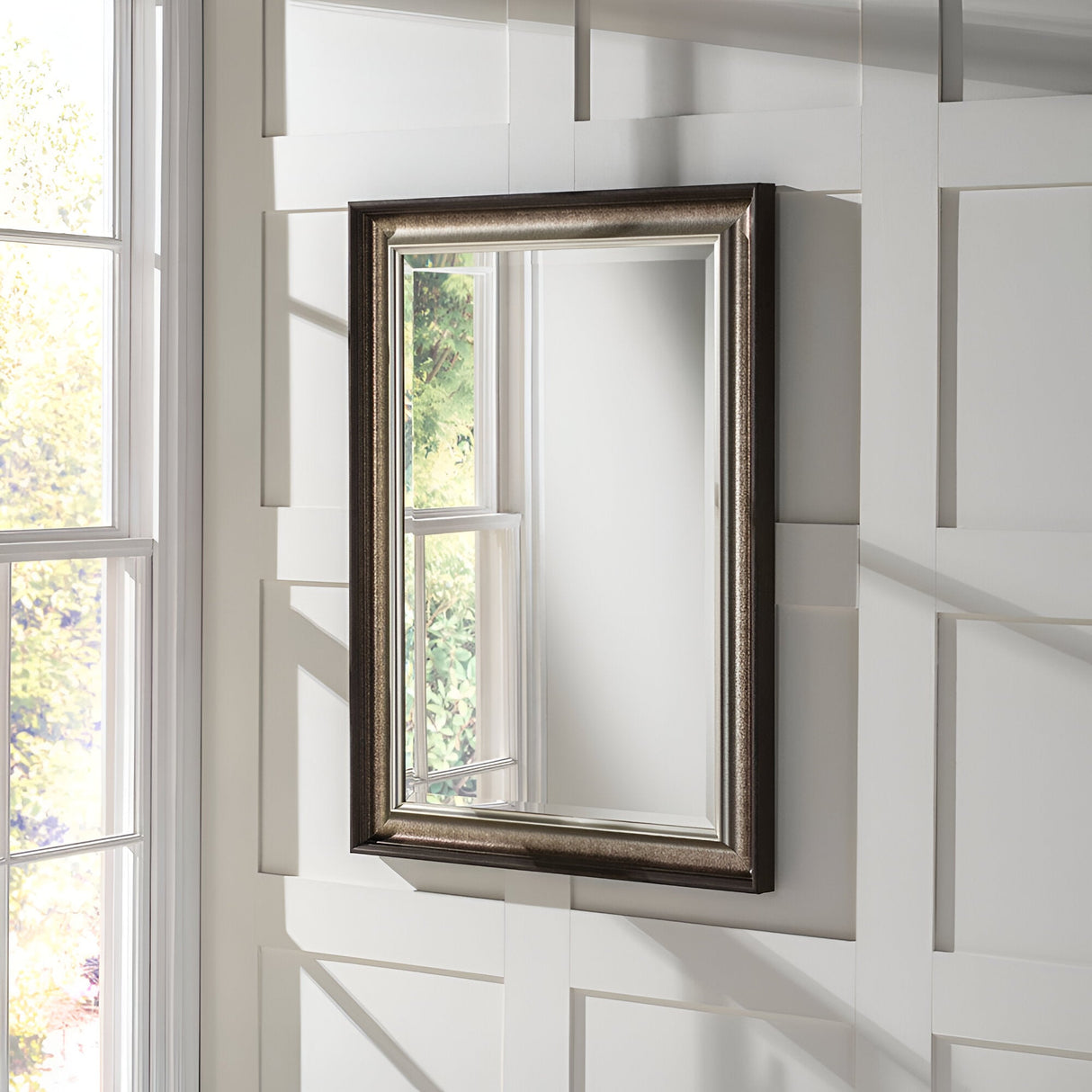 The Luxon Rectangle Wall Mirror in Antique Silver is mounted on a white paneled wall. A window beside it reflects faint sunlight and greenery, creating a serene and elegant atmosphere.
