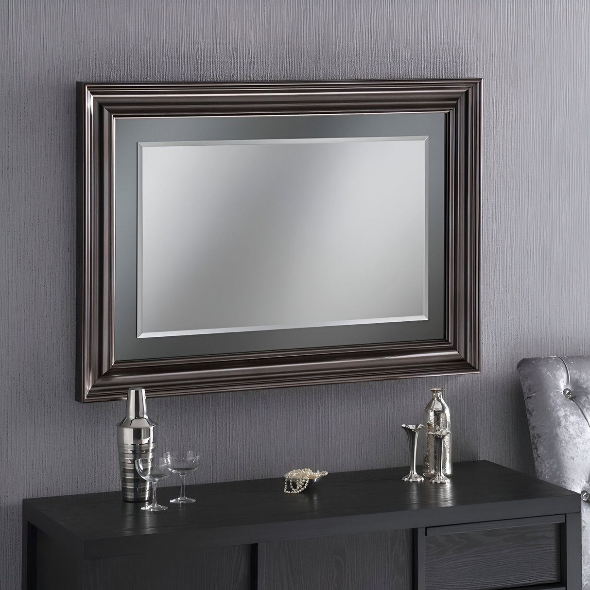 The Luxon Rectangle Wall Mirror - Silver Lacquered gracefully hangs on a gray textured wall above a black sideboard. Decorative items such as a cocktail shaker, glasses, and a small elephant figurine adorn the sideboard, adding charm to this sophisticated setting.