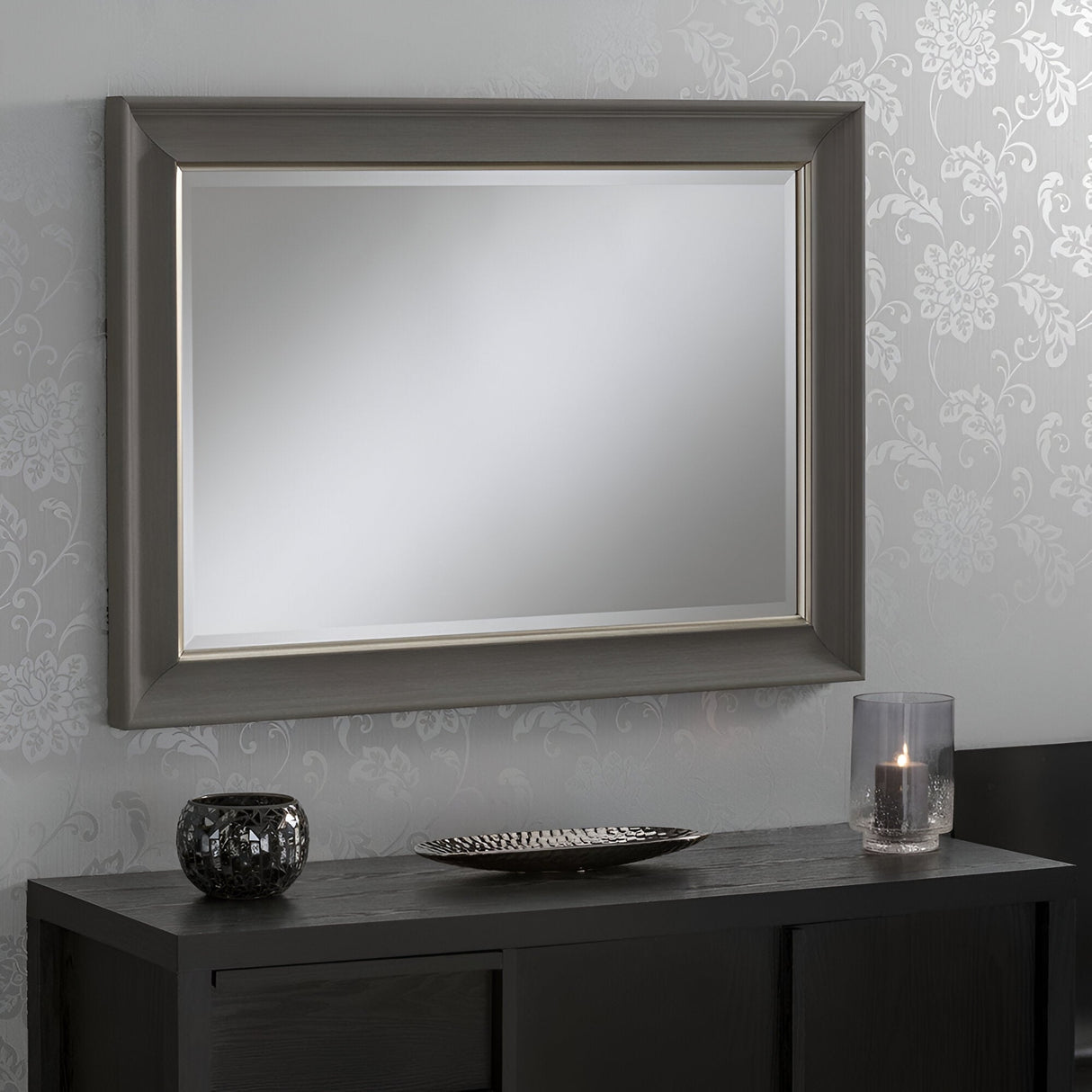 A Luxon Rectangle Wall Mirror - Dark Grey, exuding modern sophistication, is mounted on a patterned wall above a dark console table. Its frame perfectly complements the table, which is adorned with a textured candle holder, an ornamental plate, and a lit candle encased in glass.