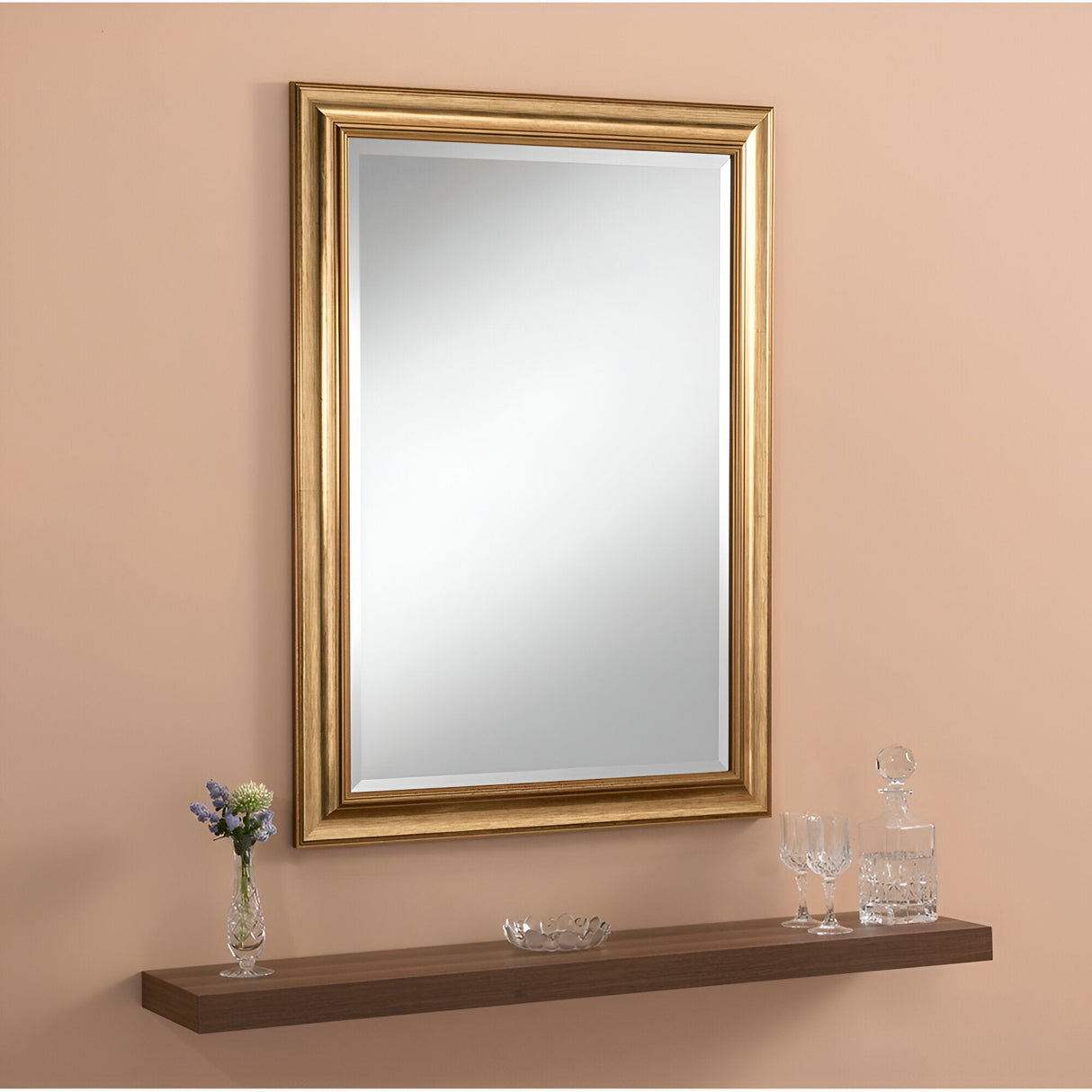 The Seraph Rectangle Wall Mirror - Gold is elegantly displayed on a beige wall above a wooden floating shelf, adding a touch of luxury to the home decor. It perfectly complements the slender vase of flowers, glass decanter with stopper, small glass bowl, and two crystal glasses.