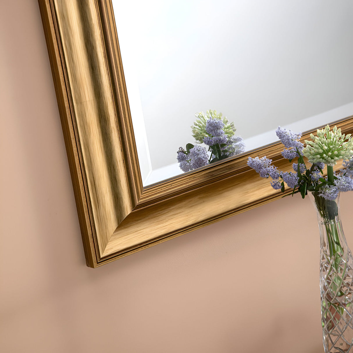 The Seraph Rectangle Wall Mirror - Gold beautifully reflects a small bouquet of lavender and green flowers. Gracefully arranged in a decorative glass vase on a beige surface, this floral display enhances luxury home decor with its elegance and natural charm.
