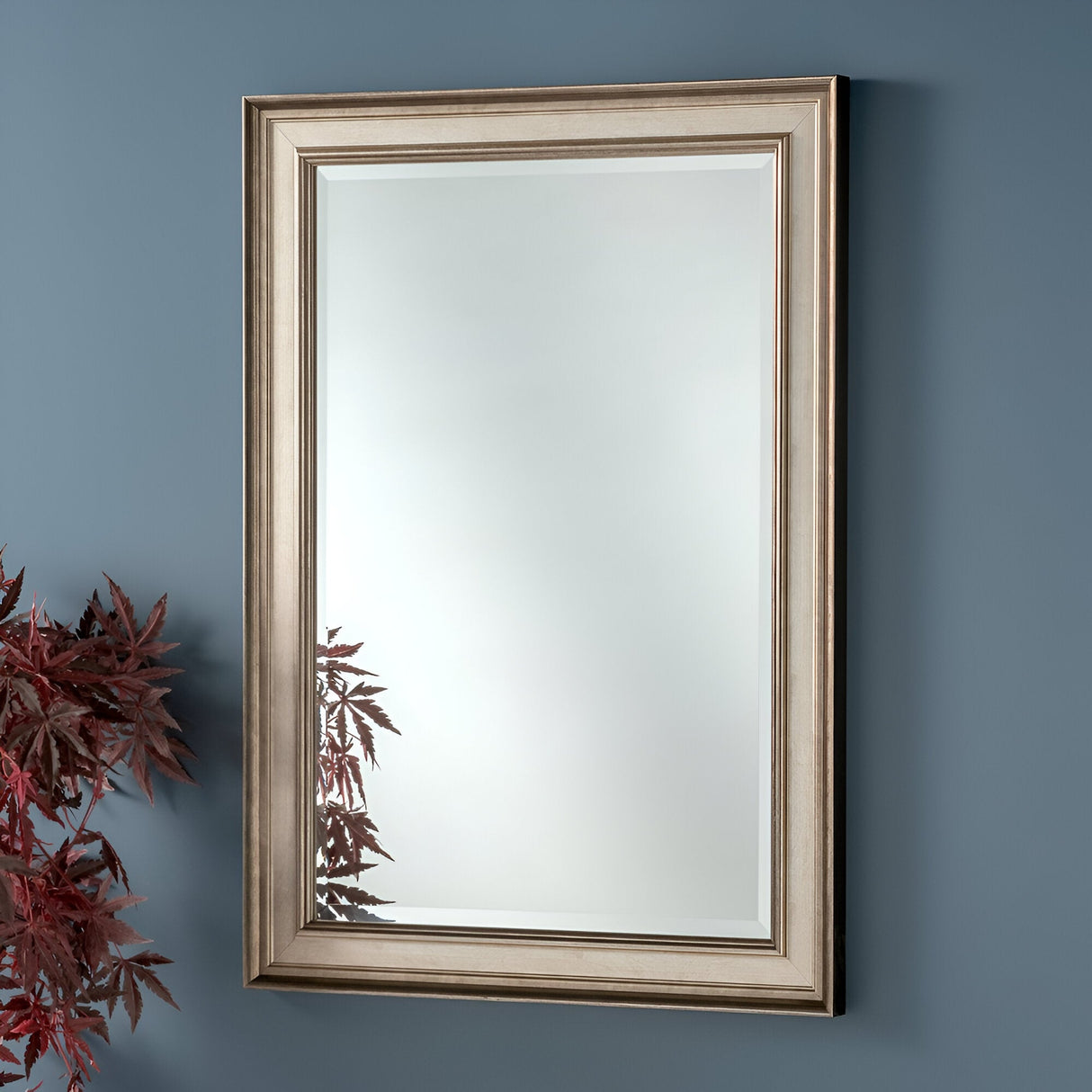 The Elysium Rectangle Wall Mirror - Silver, a marvel of modern sophistication, boasts a striking silver frame. It hangs gracefully on a blue wall, capturing the subtle hint of red foliage on the left side in its reflection.