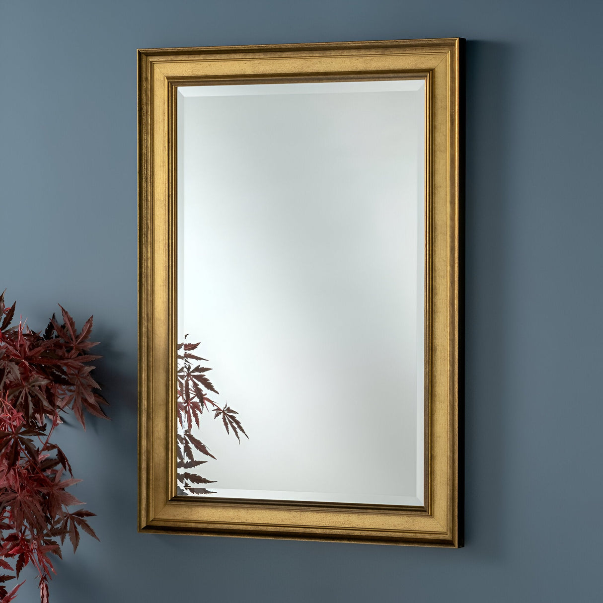 The Elysium Rectangle Wall Mirror - Gold, featuring a luxurious frame, is mounted on a blue wall. Its reflective surface captures the vibrant red foliage of a plant in the bottom left corner, enhancing the decor with an elegant touch.