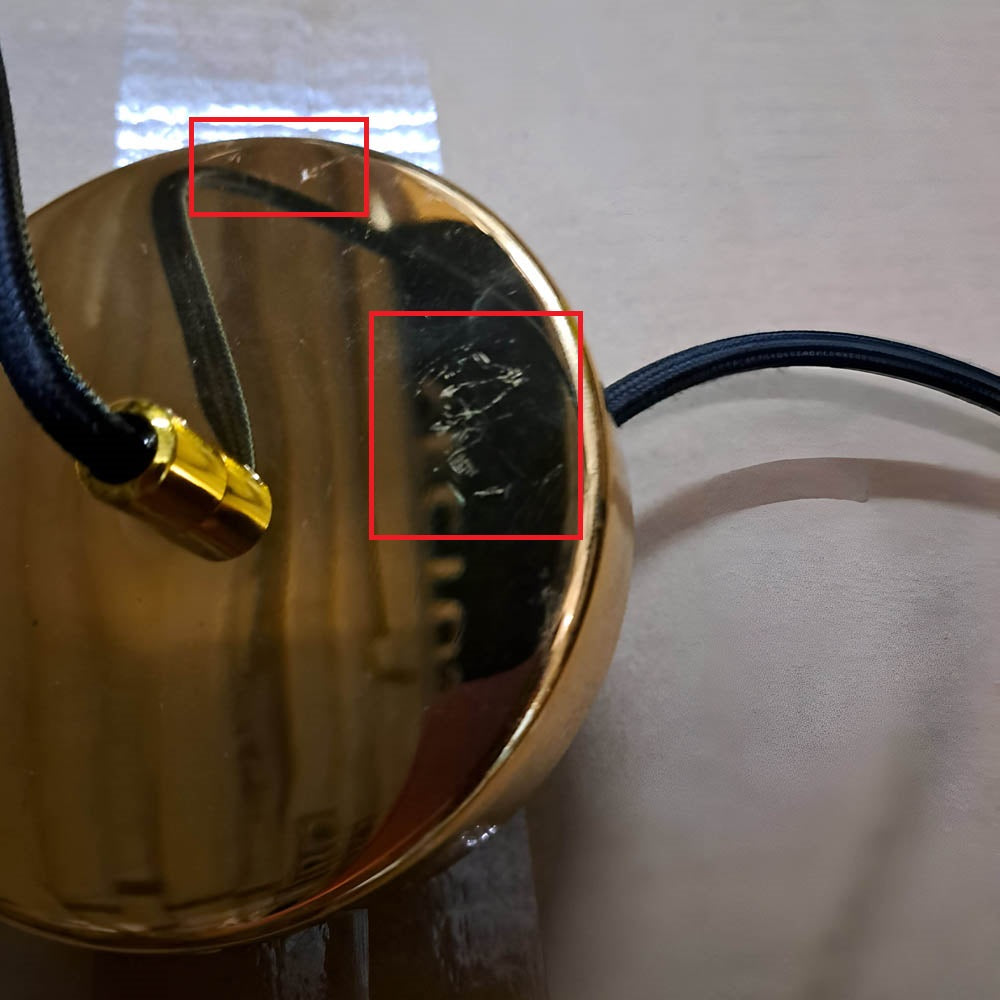 The Ex-Display Marble Pendant Light in white and gold showcases understated elegance with its shiny, circular metallic form. Visible scratches and two red square outlines enhance this luxury piece, while a black cable on the right completes the look against a neutral backdrop.