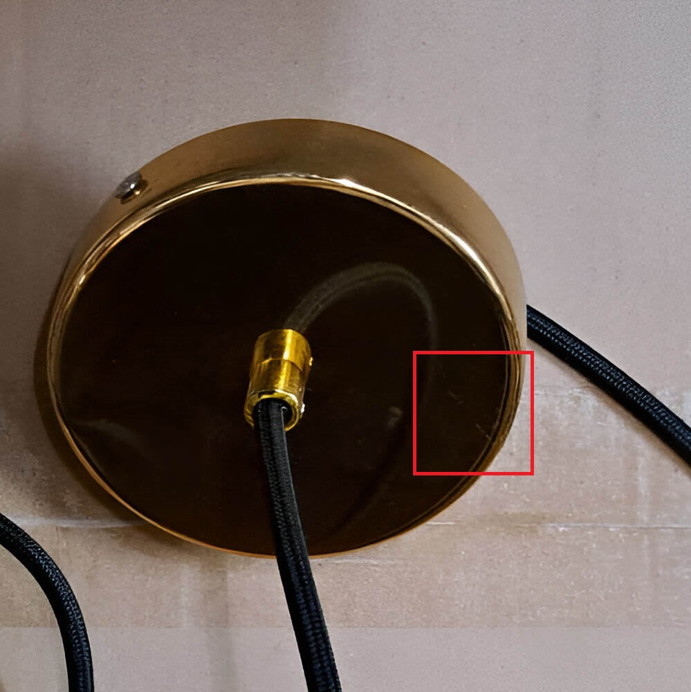 A brass ceiling fixture with a black cord partially reflects its surroundings, enhanced by a red rectangle. This luxurious lighting piece stands out against a light wall, providing an elegant touch similar to the Ex-Display Marble Pendant Light - White & Gold.