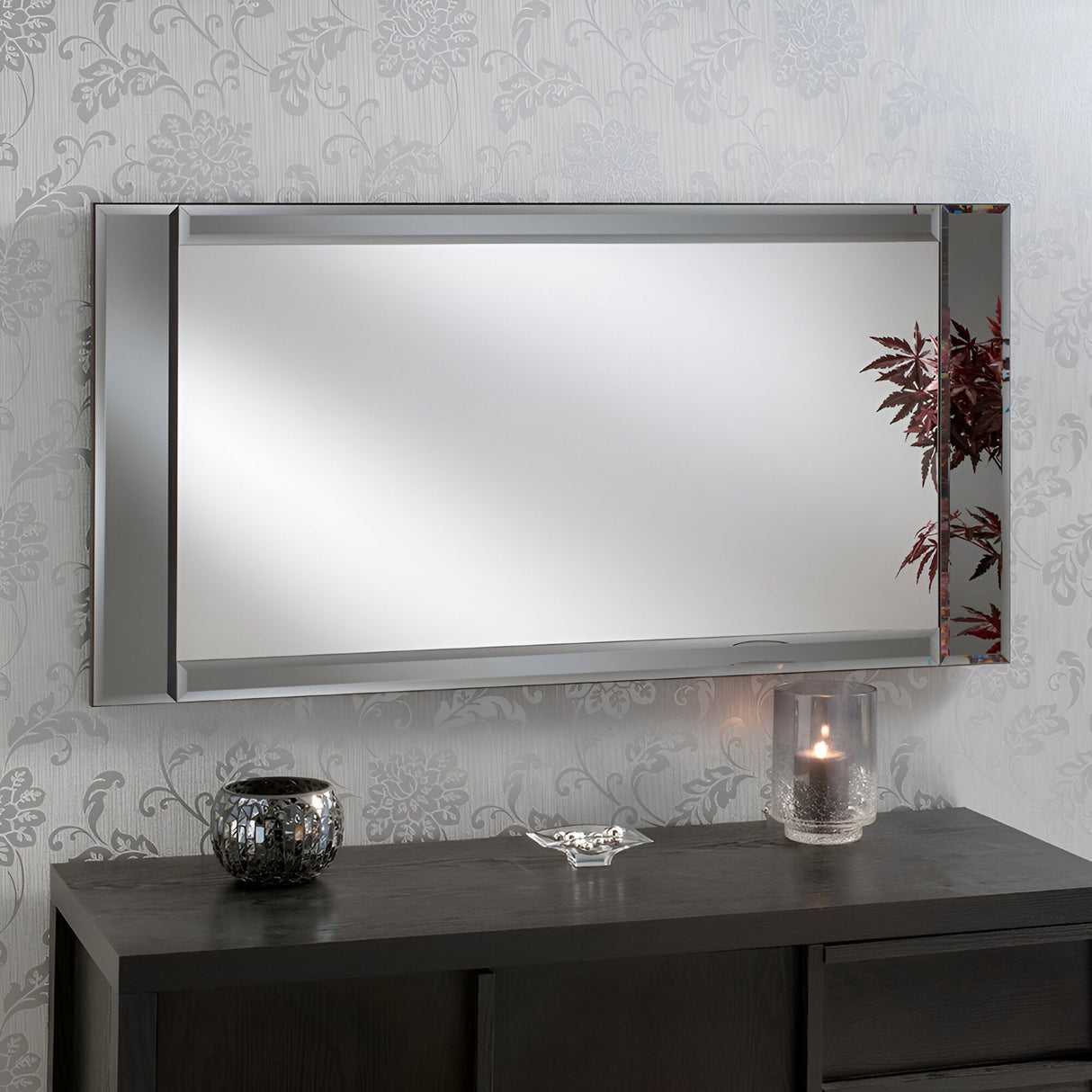 The Ambrosia Rectangle Wall Mirror (120x61cm) with a sleek wood frame on a patterned gray wall reflects the plant's red leaves. Below, a dark wooden table showcases a small decorative bowl, a textured black vase, and a glass candle holder with a lit candle, contributing to an elegant modern decor.
