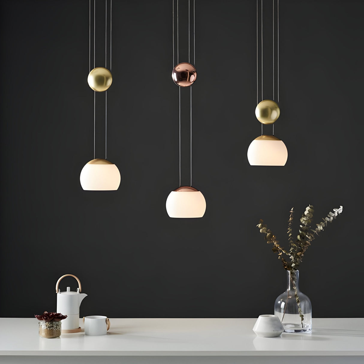 Yoyo G9 Glass Pendant Lights by Niori with opal glass shades and gold and copper counterweight spheres above a white table with teapot and vase.