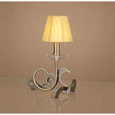 The Acanto 1 Light Table Lamp in antique brass with an amber cream shade stands on a reflective surface. Its decorative curvy base is displayed against a plain beige background.