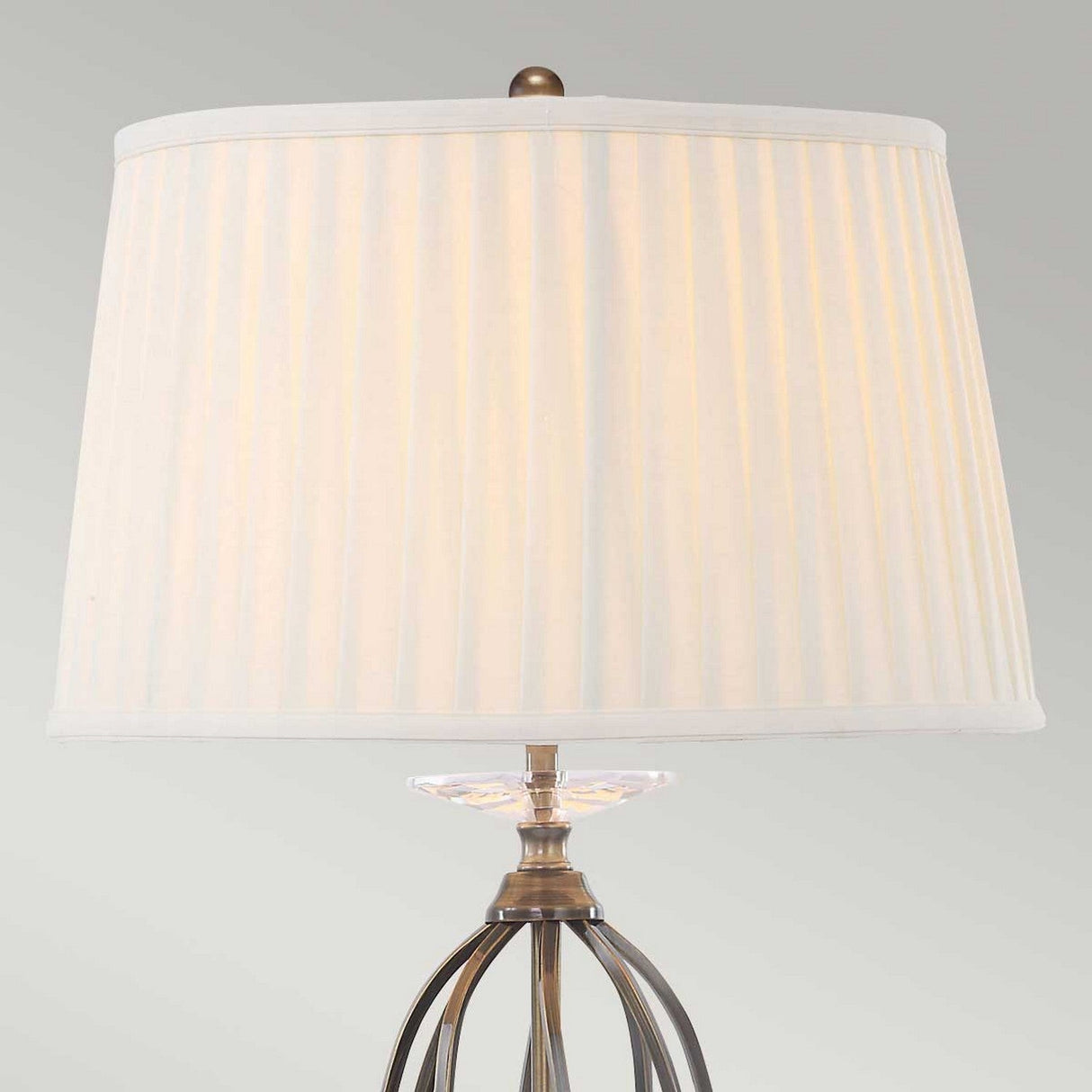 The Aegean Table Lamp - Aged Brass features a beige, pleated lampshade paired with an aged brass metal base. Its hand-forged scrolls beneath the shade introduce a transparent decorative element, enhancing its elegance against a light gray wall.