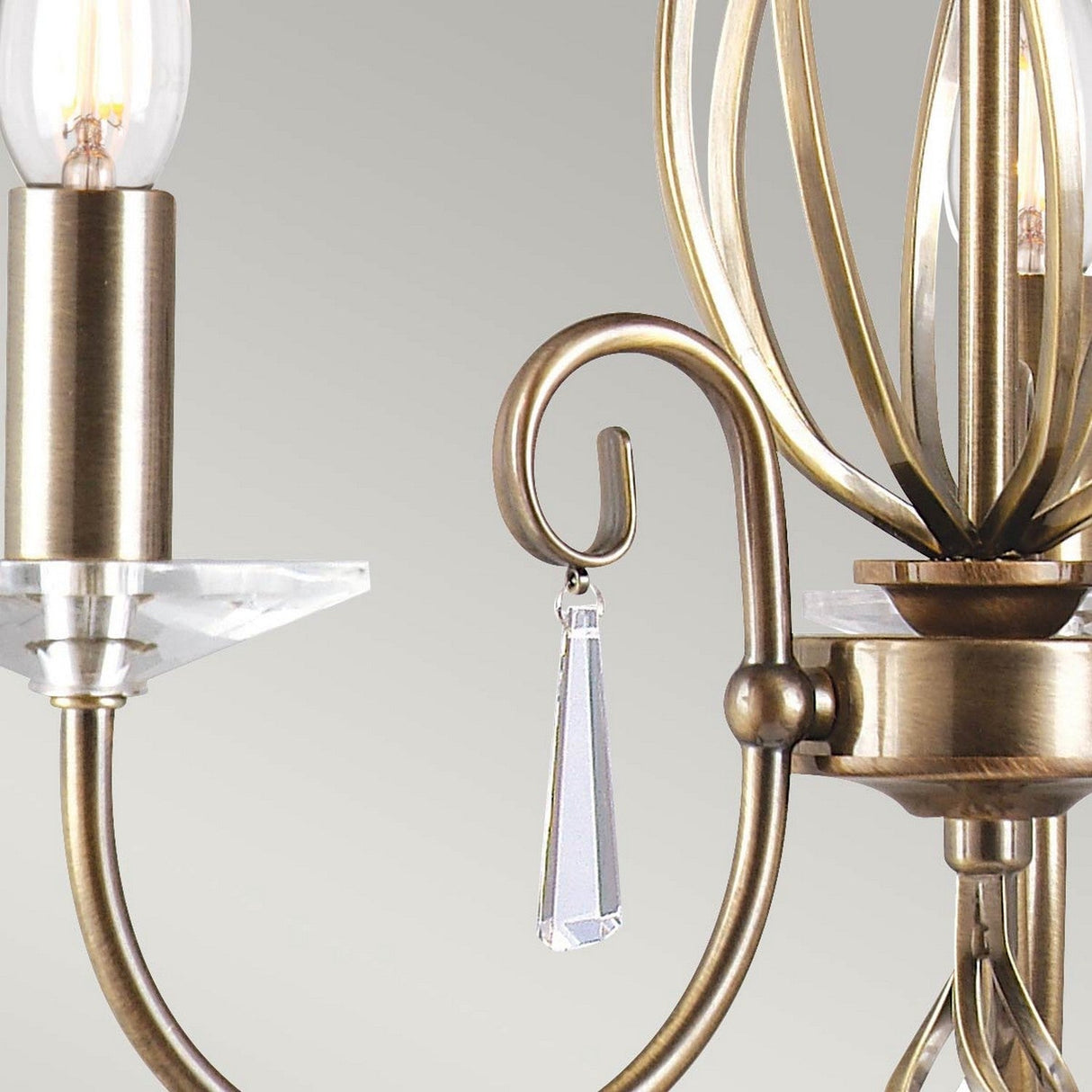 Close-up of an arm from the Aegean 3 Light Chandelier - Aged Brass, showcasing intricate detailing with a candle-like bulb and a crystal pendant. The hand-forged scrollwork evokes elegant shadows against the soft, neutral gray backdrop.