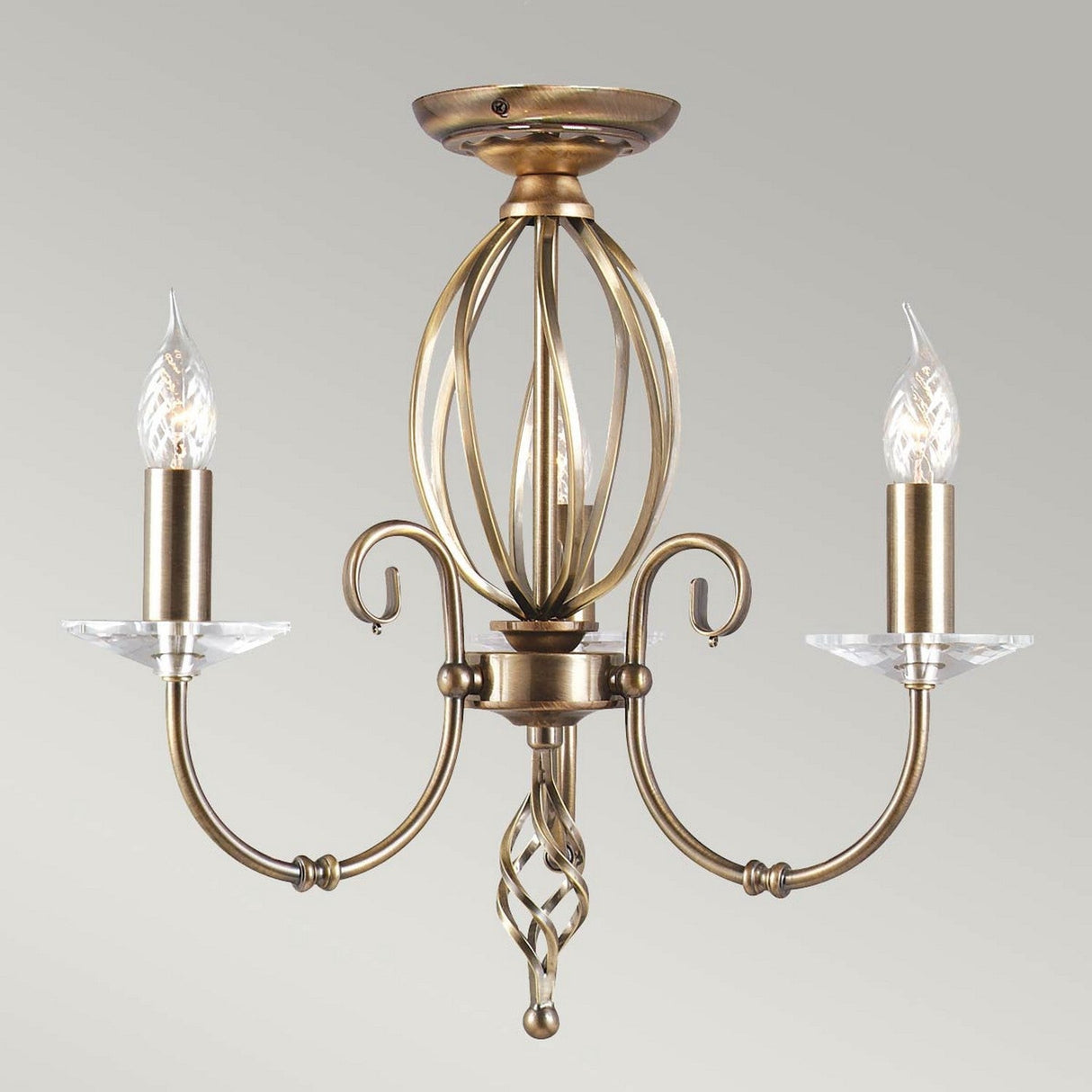 The Aegean 3 Light Chandelier in aged brass showcases a vintage design with hand-forged scrolls and a metal cage-like structure surrounding a decorative twisted centerpiece. It includes two candelabra-style bulbs that resemble candle flames.