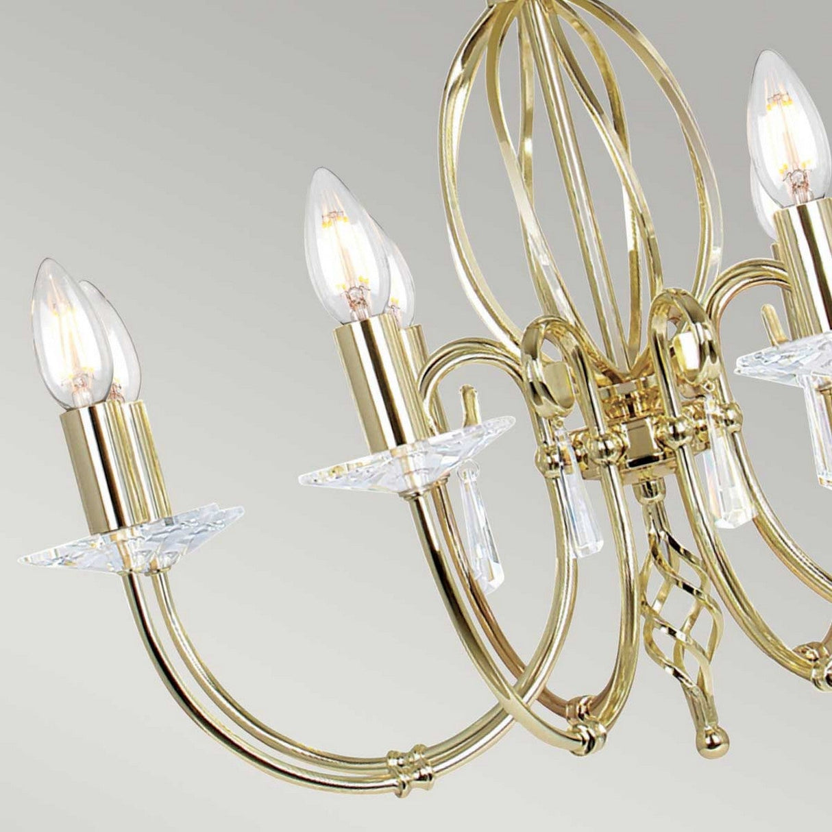 Close-up of the Aegean 8 Light Chandelier - Polished Brass, showcasing its intricately designed arms with candle-shaped light bulbs and cut glass droplets, set against a neutral background.