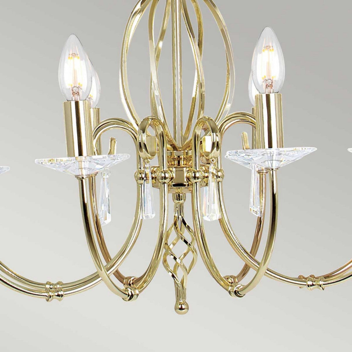 The Aegean 8 Light Chandelier - Polished Brass features an intricate design with s-shaped arms and candle-like lightbulbs. Crystal accents and cut glass droplets add elegance and sparkle as it hangs against a light gray background.