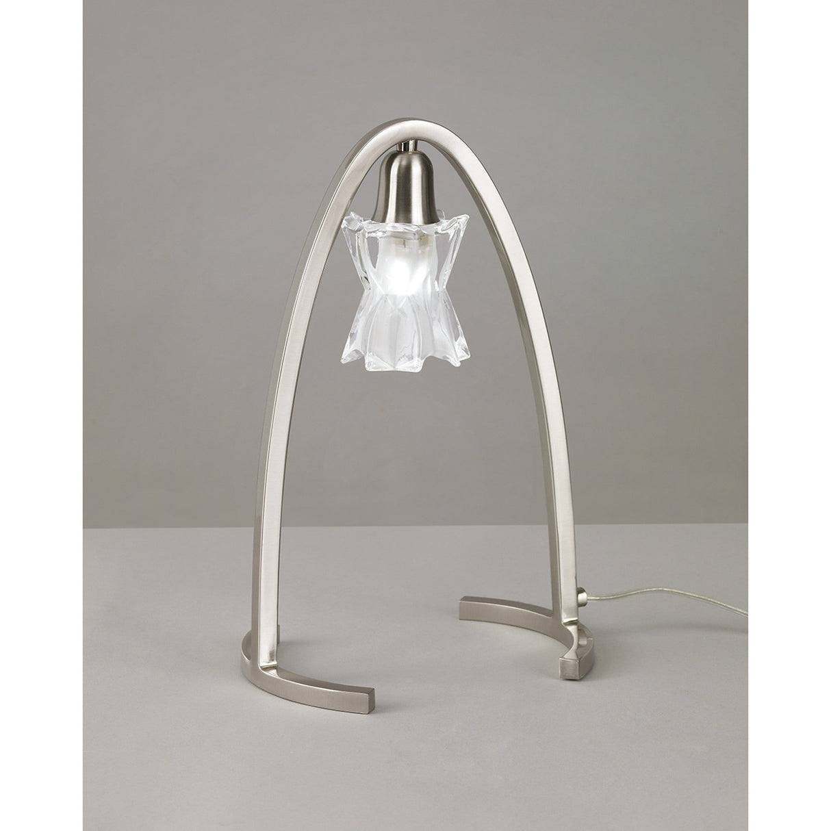 The Alaska 1 Light Table Lamp in Satin Nickel features a brushed metal arch frame and a clear, flower-shaped glass shade, shown on a neutral surface with a gray background.