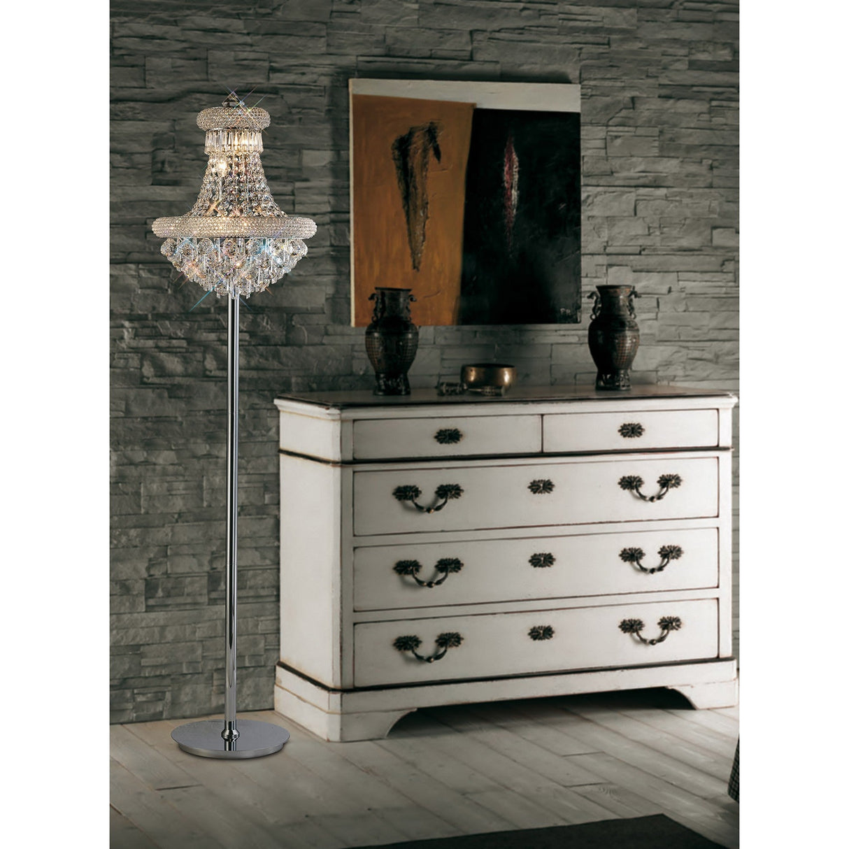 A vintage white dresser with ornate handles stands against a gray stone wall. The Alexandra 6 Light Crystal Floor Lamp in polished chrome to the left casts ambient light, highlighting two dark vases and an abstract painting atop the dresser.