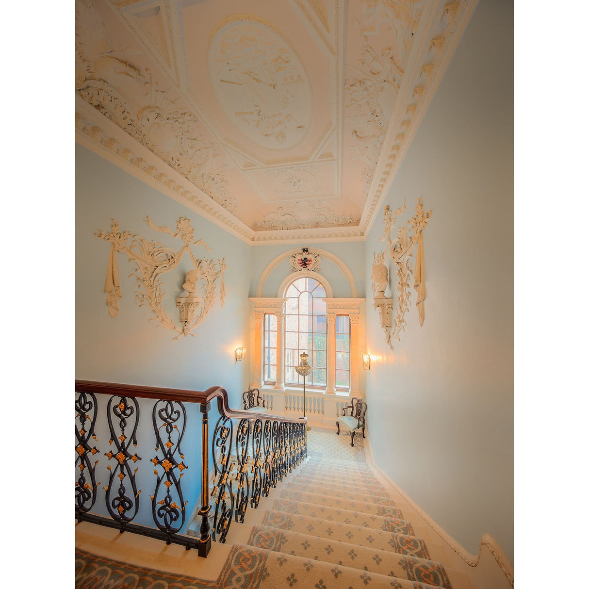 An elegant staircase with ornate railings, intricate plasterwork, patterned carpet, and a large arched window at the landing illuminated by the Alexandra 6 Light Crystal Floor Lamp in gold.