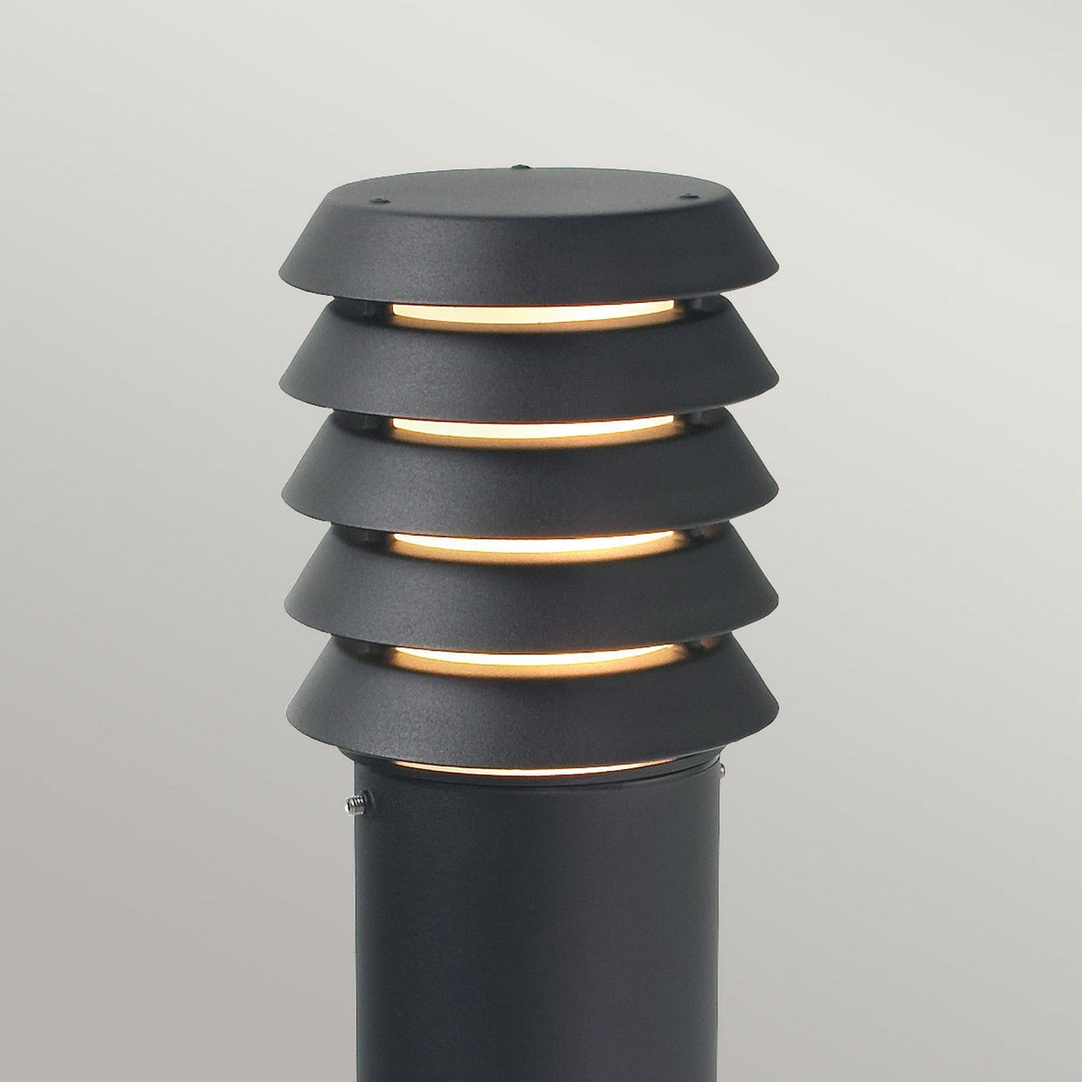 The Alta 1 Light Large Bollard - Black is a modern outdoor lamp post with a sleek, cylindrical design. It boasts an IP65 rating for durability and features horizontal, evenly spaced light fixtures that emit a warm glow, enhancing its contemporary black finish against a neutral, light grey background.