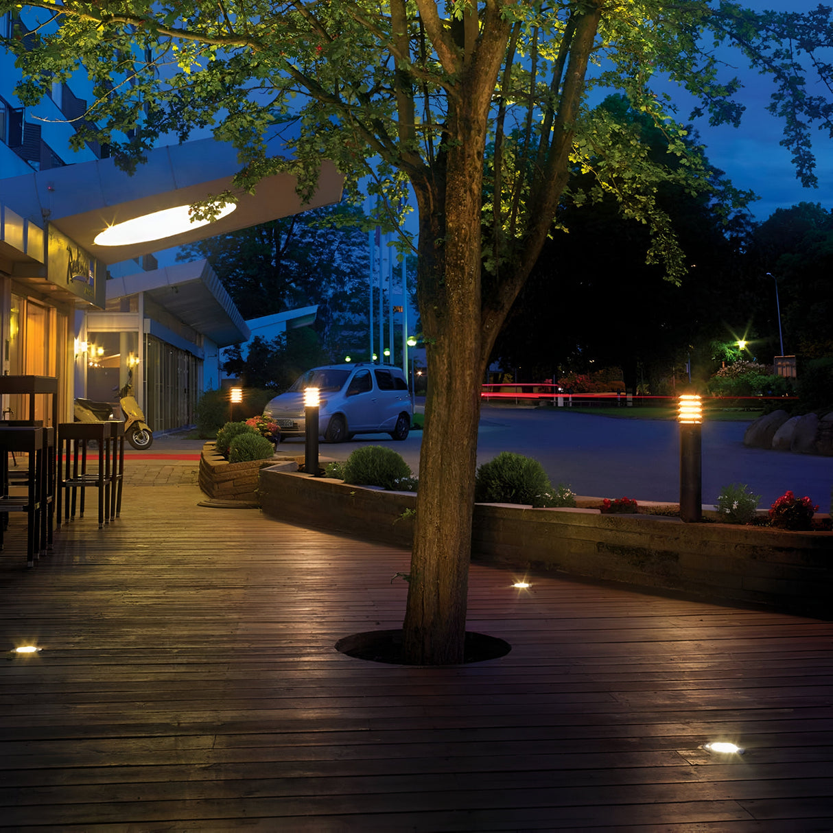 A dimly lit outdoor patio with wooden decking centers around a tree, softly illuminated by Alta 1 Light Large Bollard - Black. Its IP65 rating ensures durability as the path gleams gently. Nearby, a car is parked on the street, and a building with large windows alongside a parked scooter completes the scene.