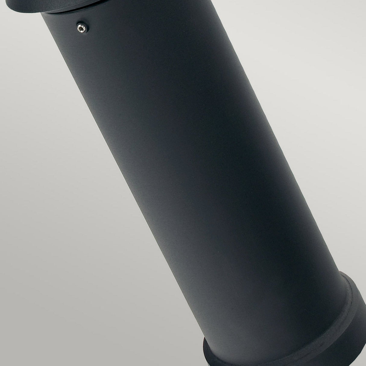 A close-up of the sleek, cylindrical Alta 1 Light Medium Bollard - Black, featuring a smooth matte finish against a neutral gray background. The perspective emphasizes one side of the cylinder, highlighting a small metal screw detail, ideal for dimmable outdoor lighting solutions.