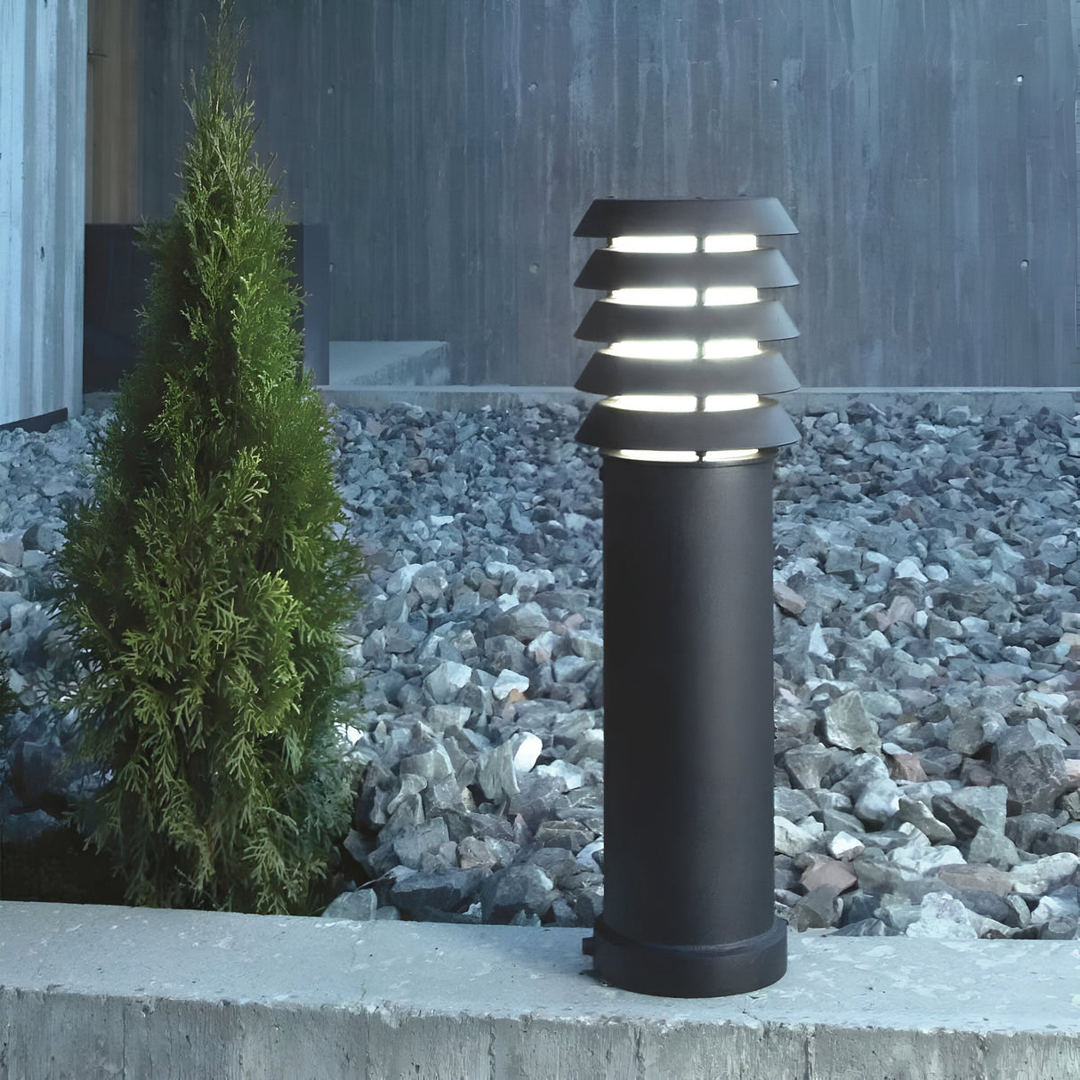 The Alta 1 Light Medium Bollard - Black, with its dimmable lighting, elegantly lights up a gravel pathway beside a small evergreen shrub and a concrete edge. Its sleek design makes it stand out against the wooden wall backdrop.