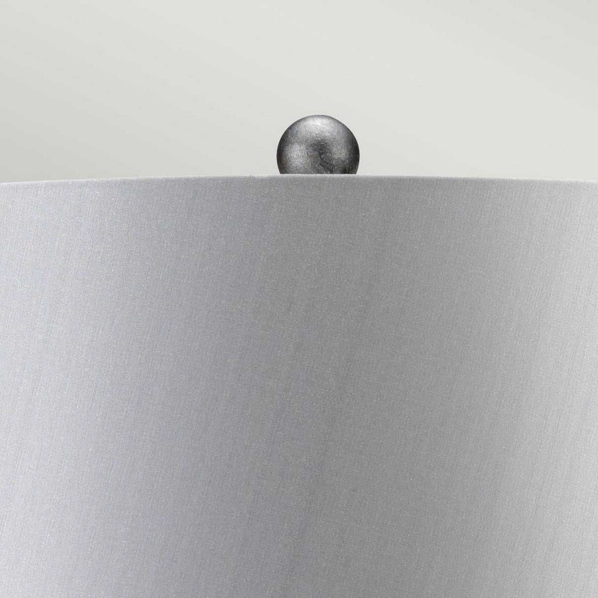 A close-up of the Amarilli 1 Light Table Lamp - Black With Silver Patina showcases its brushed metallic lampshade, topped with a small round finial, set against a light gray background. The subtle sheen highlights the handcrafted details and the charming silver patina finish.