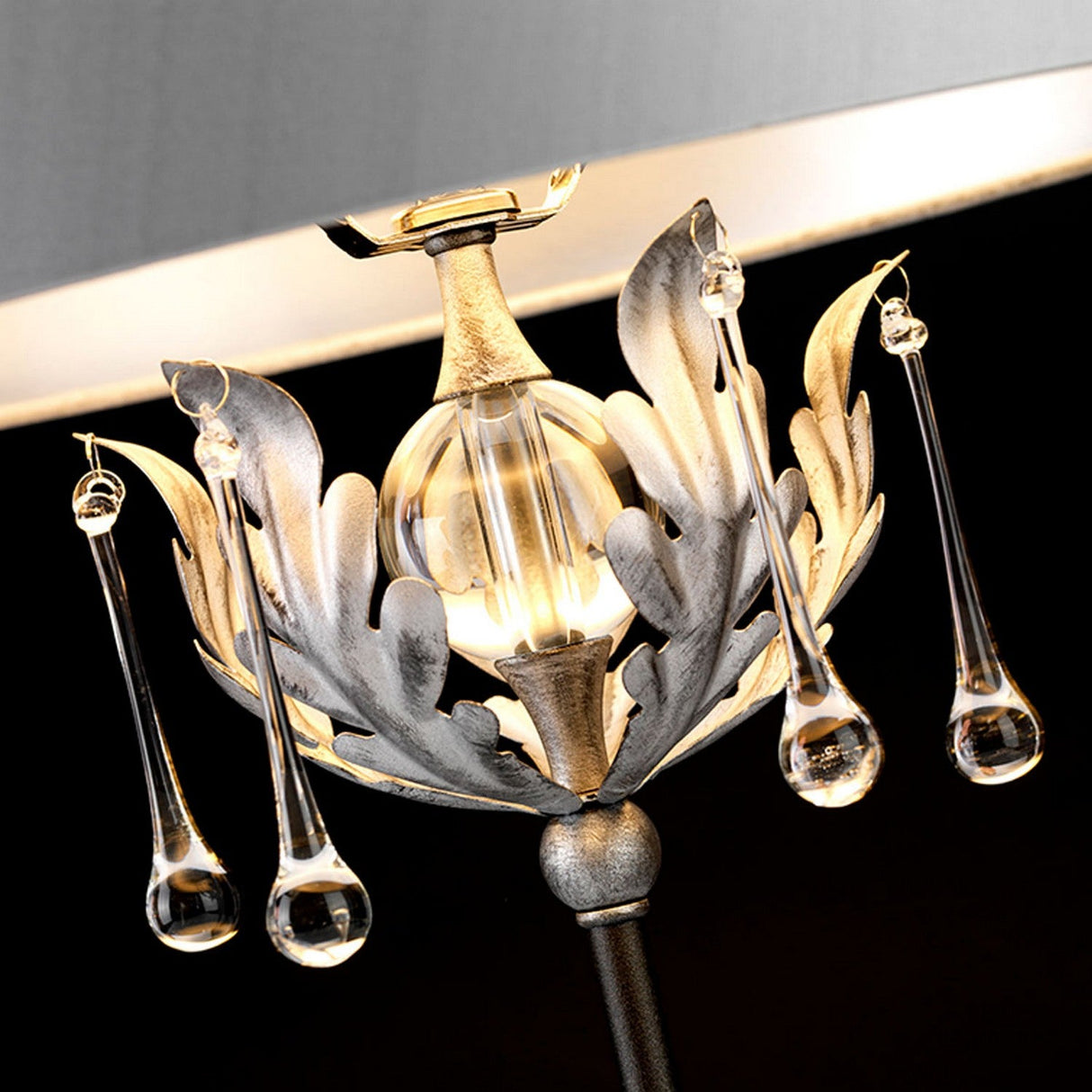 A close-up of the Amarilli 1 Light Table Lamp - Black With Silver Patina highlights its intricate fixture featuring a spherical bulb encircled by leaf-like metal petals. Exquisite handcrafted details include crystal teardrop ornaments, and the piece is topped with a circular lampshade, making it a stunning decorative statement.