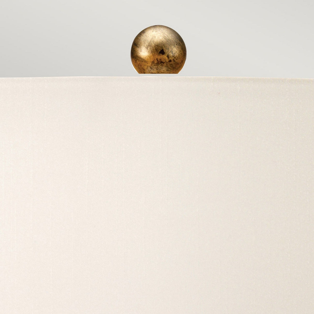 A close-up of the Amarilli 1 Light Table Lamp, showcasing its ivory lampshade and decorative gold sphere finial on top, adorned with a bronze base featuring a subtle gold patina finish. The soft gray background enhances the minimalist and elegant ambiance.