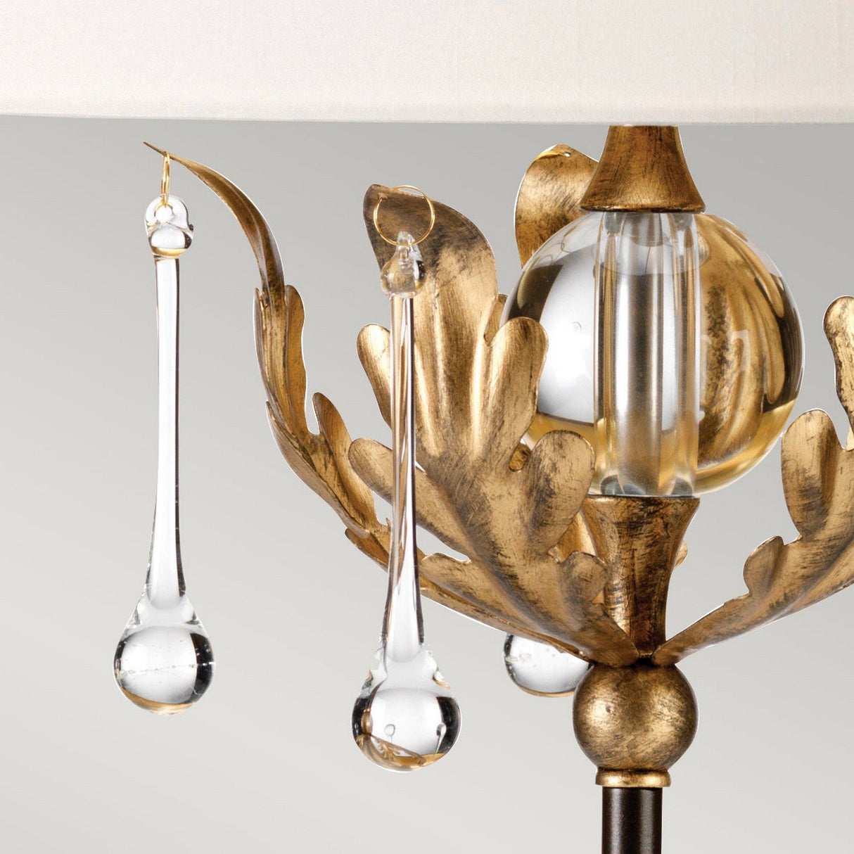 Here's a close-up of the Amarilli 1 Light Table Lamp, featuring an elegant bronze structure with a gold patina finish and clear glass accents. The design includes two glass teardrop shapes elegantly hanging from ornate leaves, surrounding a spherical glass centerpiece beneath an ivory shade. This handmade piece casts ambient light beautifully.