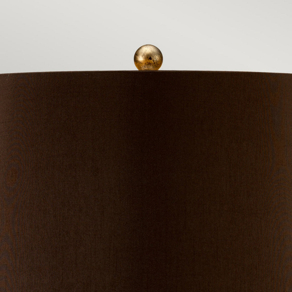 The Amarilli 1 Light Table Lamp is beautifully showcased with its brown cylindrical lampshade featuring a smooth texture. At the top, it is adorned by a small, round finial in bronze with a gold patina, adding elegance against a neutral backdrop.