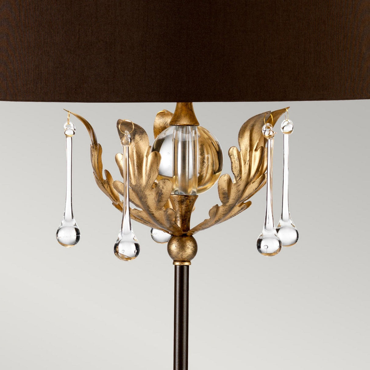 The Amarilli 1 Light Table Lamp - Bronze With Gold Patina & Brown Shade is an elegant, handmade lamp featuring a dark brown shade and a decorative stem adorned with gold leaf-like patterns. Luxurious bronze with gold patina ornaments hang from the stem against a soft gray background.